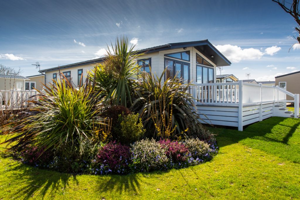 Large caravan with decking sits next to a grand rockery filled with shrubs of multi height and colour