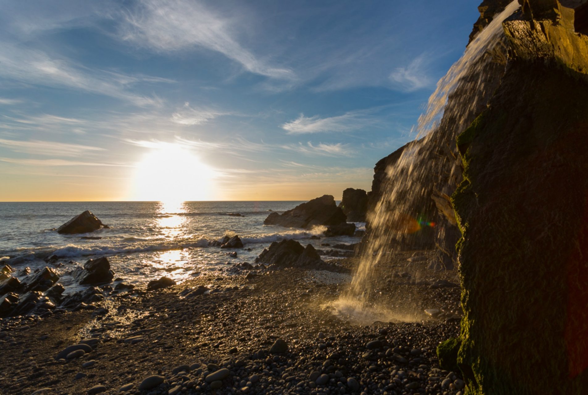 Sandymouth beach with rocks and sand at sunset. Waterfall from the cliffs with sunflare
