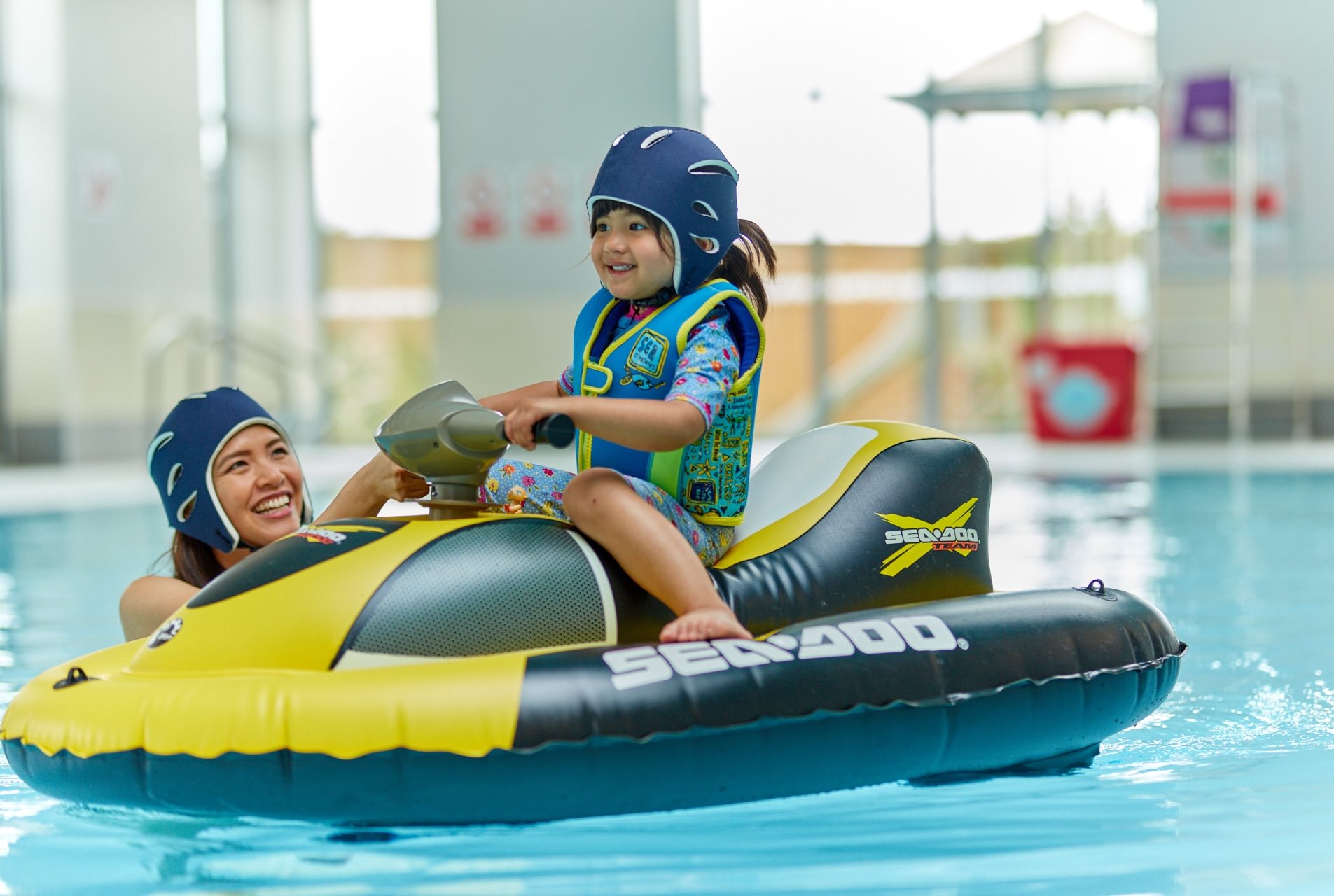 little girl in safety helmet and jacket rides an inflatable jetski in an indoor pool/ Mum watches close by