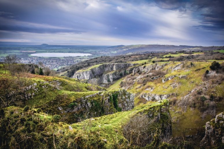 dramatic countryside under stormy grey-blue skies. Cargs and cliffs in teh foregound, witha large round lake in the cetnre. A large hillock stands on an otherwise flat horizon, two people can be seen on a cliffedge