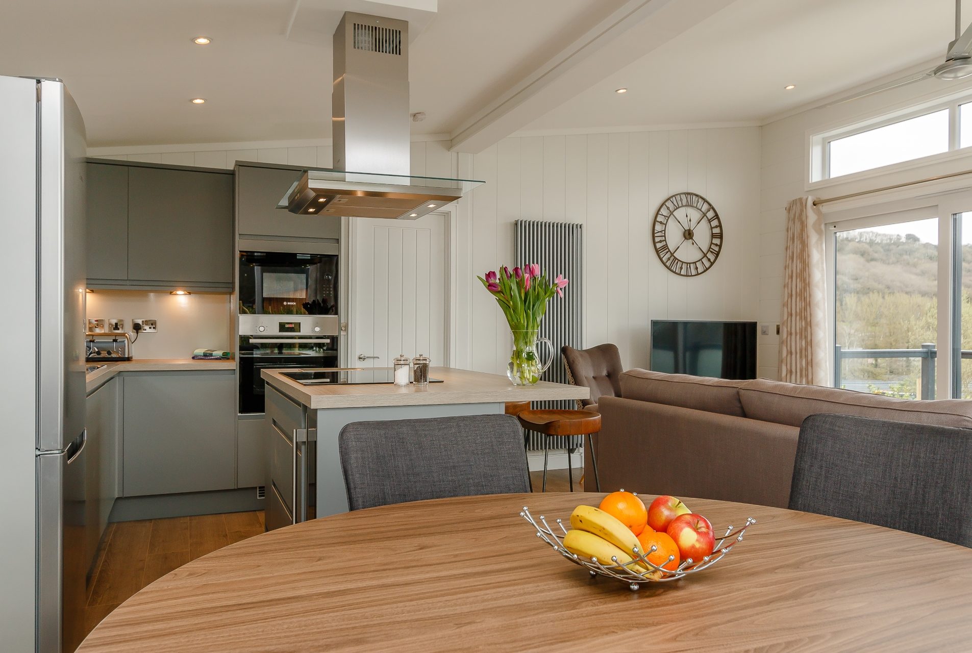 A modern open-plan living area with green fitted kitchen, large dining table and the back of a beige couch. a large clock is on the wall and a glimpse of countryside can be seen through the patio doors