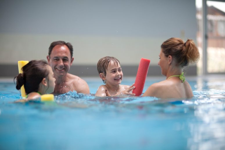 family of four play with foam pool noodles on an indoor swimming pool