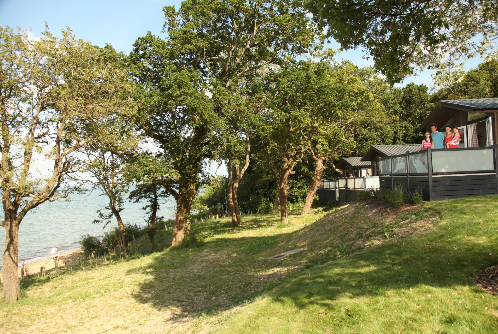 family on the decking of a lodge surrounded by greenery, with the sea to the left