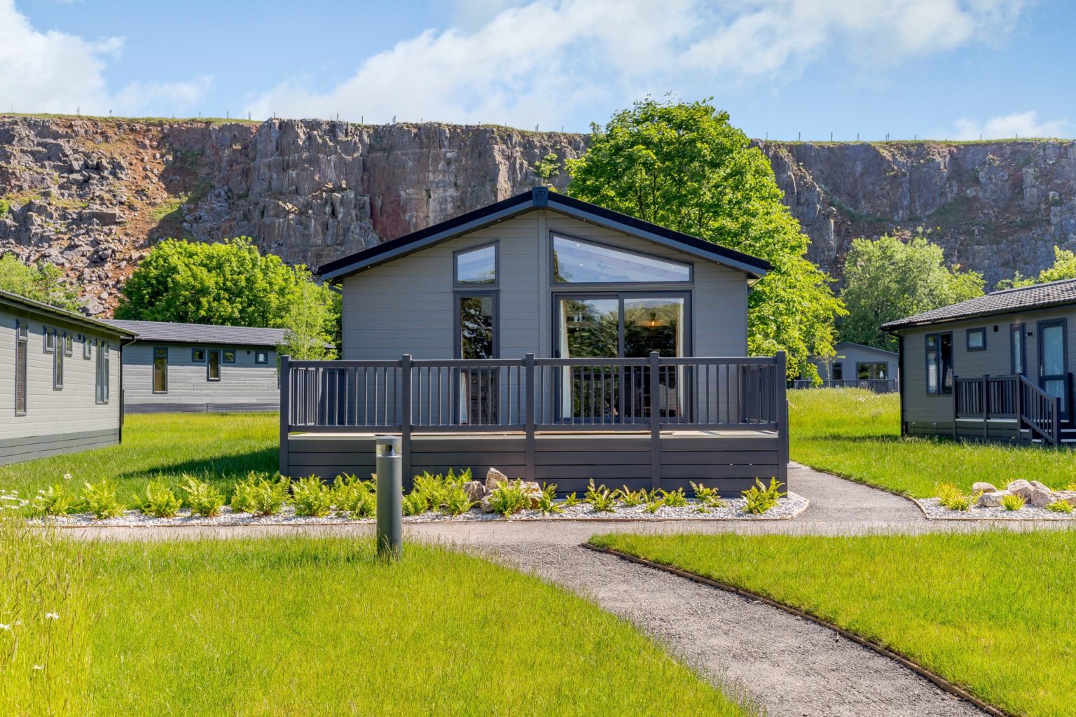 Row of grey clad lodges with deckings. Surrounded by green lawns and quarry face backdrop