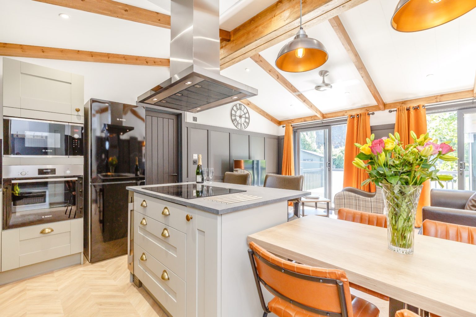 Modern open-plan living area with grey kitchen units, gloss black fridge, pendant lights, orange dining chairs and grey leather sofa