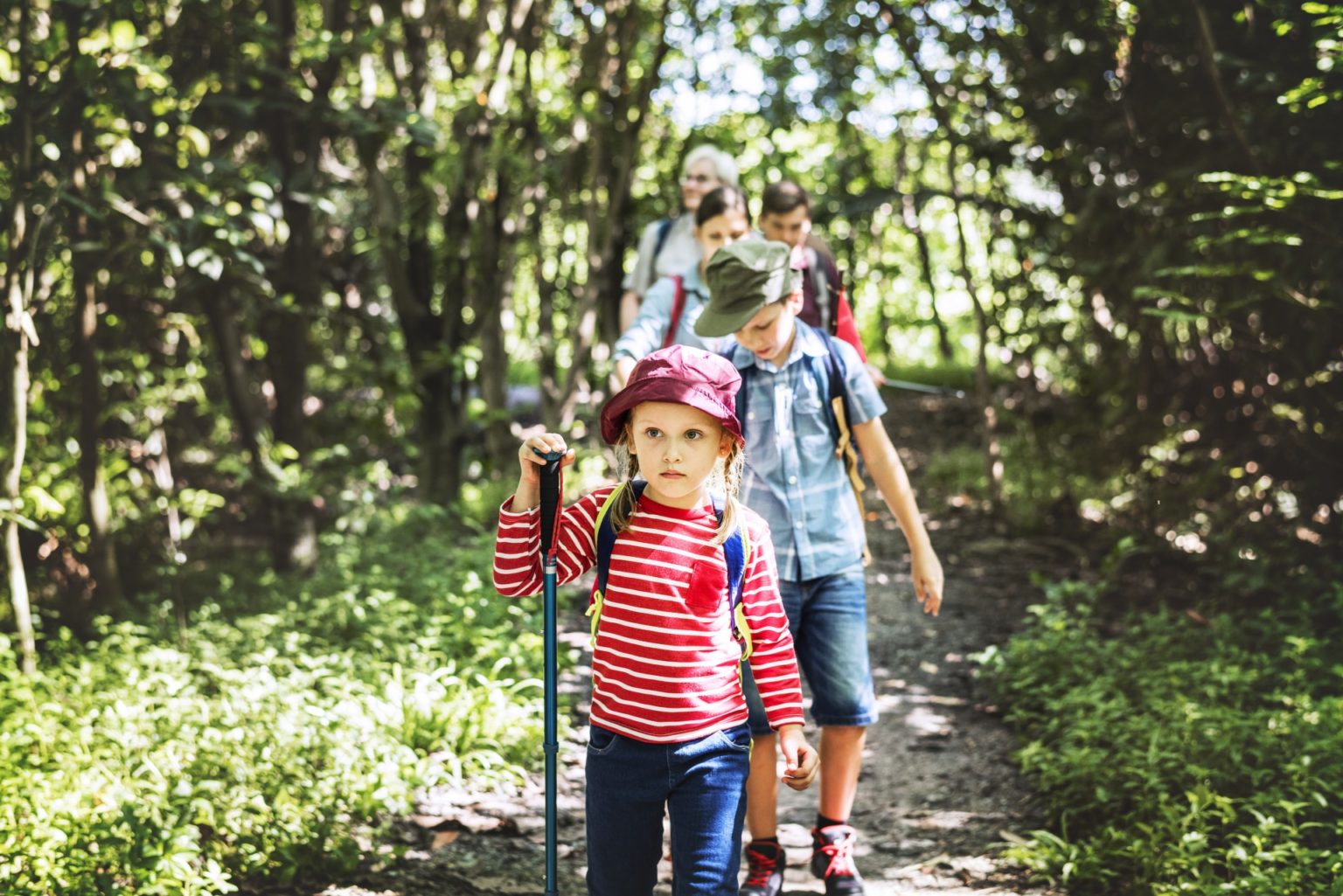 Family of four walk towards the camera with backpacks and hiking sticks, the little girl at the front wears red striped top
