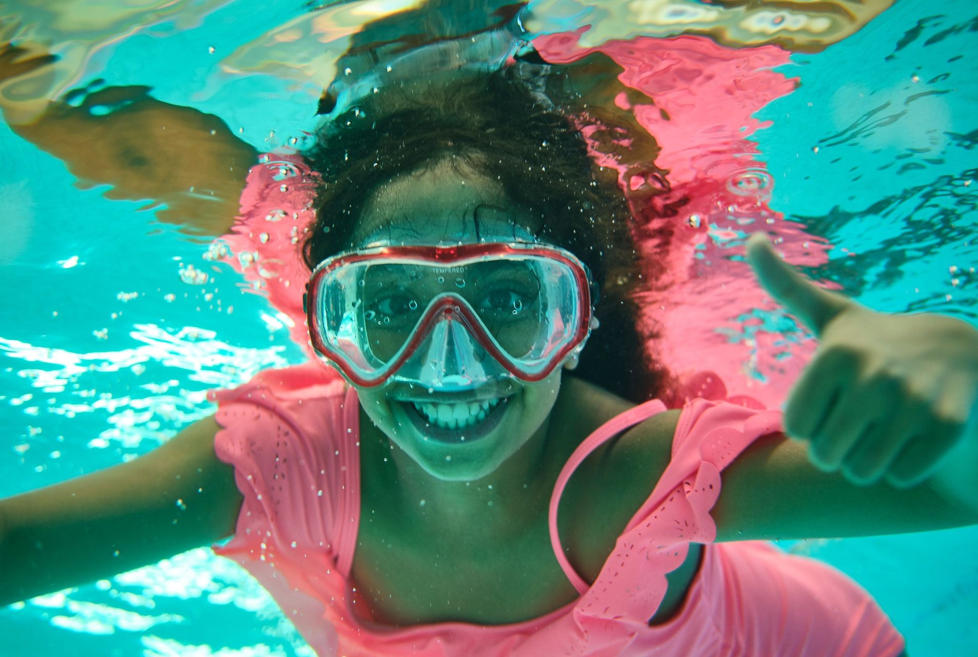 Gilrl with pink swimming costume and goggles smiles under water
