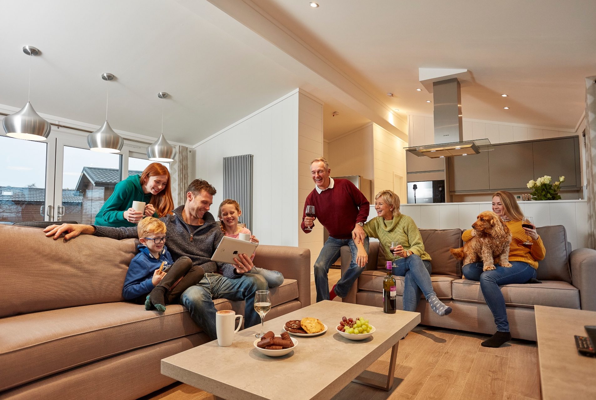 Large family relax in an open plan living area. they drink wine, eat snack, cuddle a dog and gather round a tablet