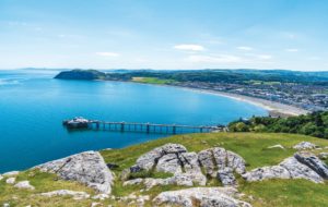 Llandudno is seen from the Great Orme. rocks and grass in the foreground, a large pier stretches into blue sea with the victorian sea front buildings seen to the right