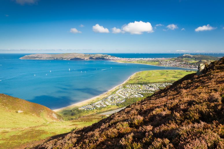 The Great Orme can be seen from Conwy Mountian. We look down on blue sea, the tide is in and yachts can be seen. We see a caravan park (aberconw) from a birds-eye-view