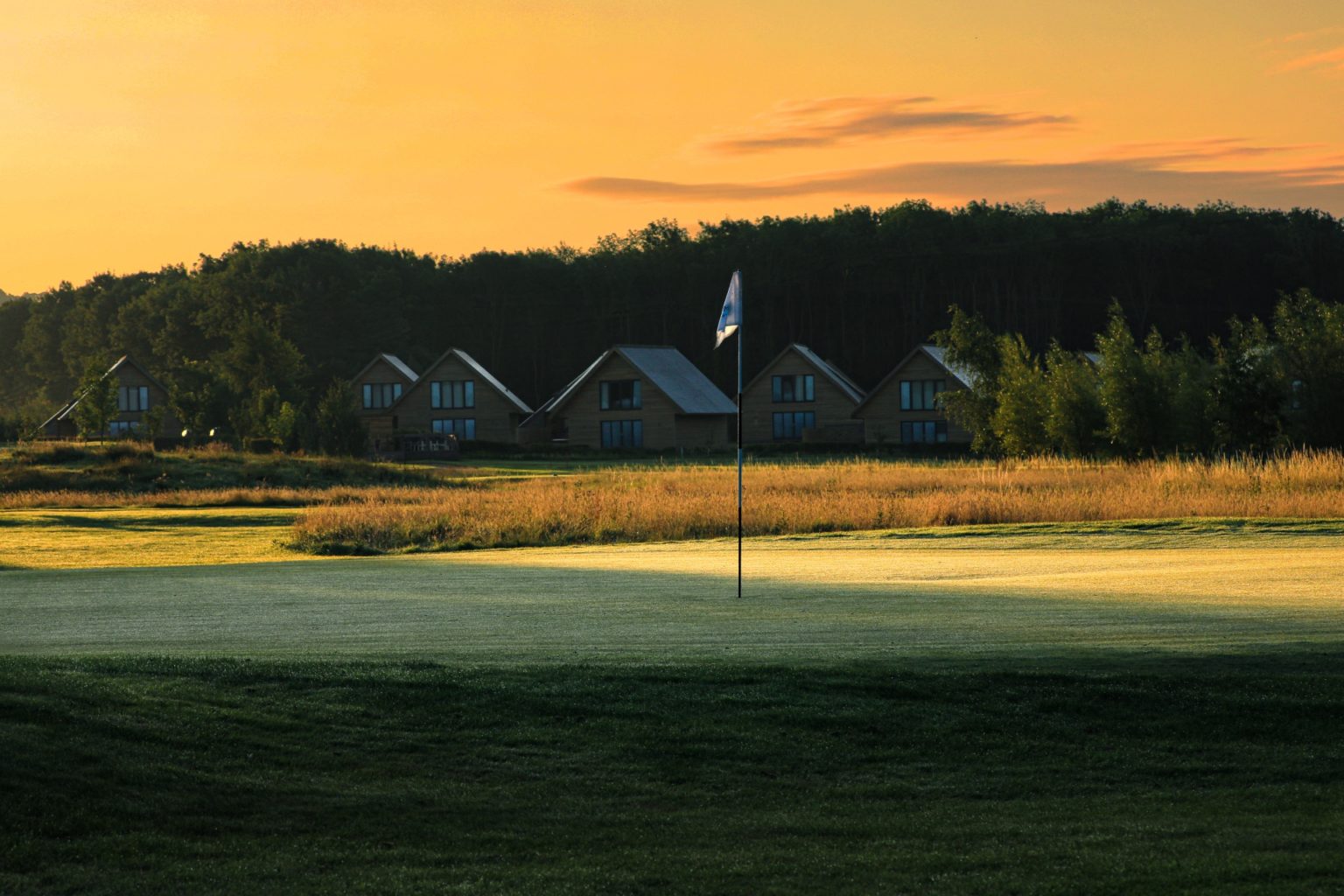 Row of lodges with pitched roofs , the sun is rising and there's a light frost in the foreground on the golf course, with flag